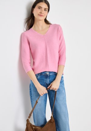 Frau trägt ein rosa Dreiviertelarmshirt und blaue Jeans, hält eine braune Ledertasche mit beiden Händen vor sich.