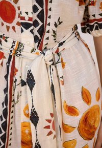 Weißes Kleid mit bunten Blumen- und Sonnenmustern. Verfügt über kurze Ärmel, einen Knopfverschluss und einen gebundenen Taillengürtel. Texturiertes, leichtes Material.
