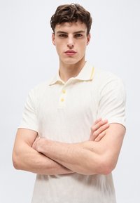 Νέος άνδρας με κοντά καστανά σγουρά μαλλιά, που φοράει λευκό κοντομάνικο πόλο με κίτρινα κουμπιά, σταυρωμένα τα χέρια μπροστά σε απλό φόντο.