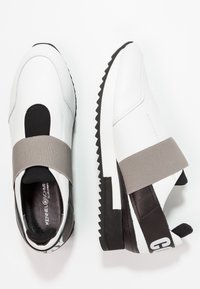 Vit sneaker med elastisk grå rem, svart häl och texturerad sula. Den släta läderöverdelen har kontrasterande paneler och varumärke.