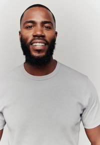 Harmaa ribbineulottu T-paita, jossa pyöreä pääntie, yllä hymyilevä tumma partainen mies. Tausta on yksivärinen valkoinen.