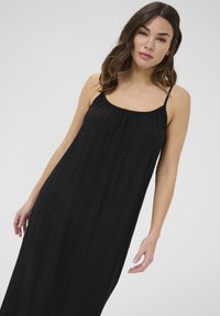 Vestido negro sin mangas con un corte holgado, escote recogido y tirantes delgados. Tejido ligero y texturizado que cae hasta el suelo.