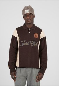 Polo a maglia color block marrone e crema con maniche lunghe, dotato di logo ricamato e testo blu. Abbinato a pantaloni grigi a pieghe e un berretto.