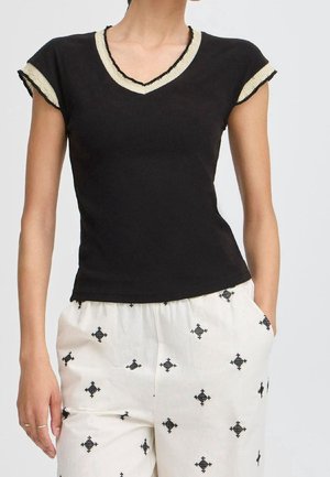 Černé elastické tričko s krátkými rukávy a výstřihem do V, ozdobené krémovými a černými lemy. Nošeno s bílými kalhotami zdobenými černými diamantovými vzory.