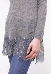 Pull gris avec une texture douce, doté d'un ourlet en dentelle d'un gris plus clair, et d'une petite étiquette logo sur le côté. Porté avec un jean bleu.
