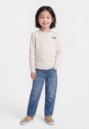 Usmievajúce sa dieťa s vlasmi po plecia, oblečené v svetlobéžovom tričku s dlhými rukávmi, modrých džínsoch a béžových slip-on topánkach, stojace na bielom pozadí.