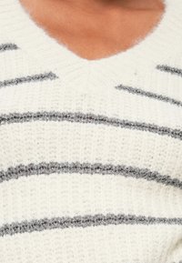 Pull en maille blanche avec un col en V et des rayures grises horizontales, présentant une texture douce et un aspect douillet.