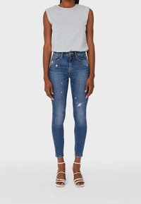Mujer con top sin mangas gris, jeans de tiro alto ajustados desgastados azules y sandalias de tacón blancas de tiras, de pie sobre fondo blanco.