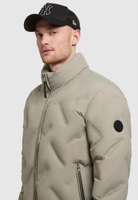 Uomo che indossa un giubbotto beige imbottito con zip e colletto alto, insieme a un cappellino nero con rete sul retro e logo bianco.