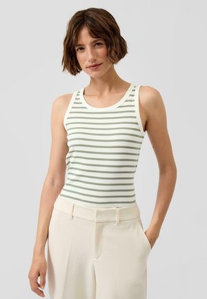 Młoda kobieta o krótkich brązowych włosach, ubrana w biały i szary poziomo paskowany bezrękawnik oraz kremowe spodnie, stoi z jedną ręką w kieszeni.