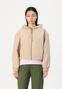 Veste à capuche beige coupée avec une fermeture éclair à l'avant, deux poches et des poignets élastiques, superposée à un haut texturé rose clair et à un pantalon plissé vert.