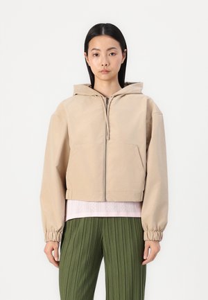 Geknipte beige hoodie met een rits aan de voorkant, twee zakken en elastische manchetten, gelaagd over een lichtroze gestructureerd topje en groene geplooide broek.
