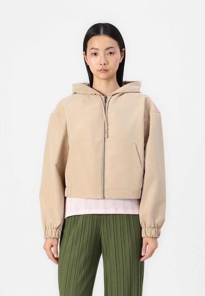 Veste à capuche beige coupée avec une fermeture éclair à l'avant, deux poches et des poignets élastiques, superposée à un haut texturé rose clair et à un pantalon plissé vert.
