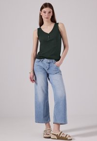 Canotta verde scuro con scollatura a V e due bottoni, abbinata a jeans a gamba larga blu chiaro e sandali marroni pelosi.