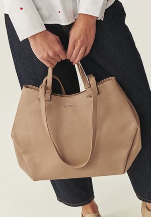 Kéz tart egy bézs bőrből készült, dupla fogantyús szatyrat, sötét farmerrel és kis piros szívmintás fehér inggel viselve.