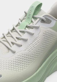 Chaussure de course en maille blanche et vert clair, avec des lacets blancs, une semelle texturée, et une boucle en tissu verte sur la languette avec un texte imprimé.