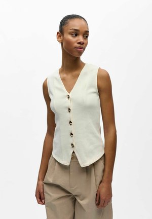 Jauna moteris dėvi be rankovių kremine liemene su sagomis ir smėlinės spalvos aukšto liemens kelnes, žvelgia į kairę, stovėdama prieš baltą foną.