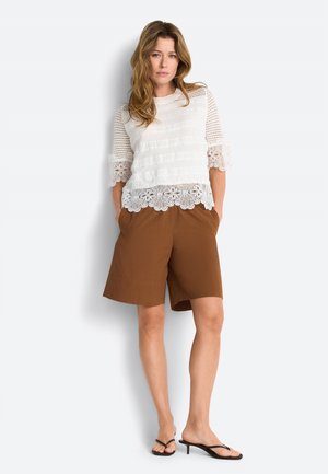 Frau steht mit den Händen in den Taschen, trägt ein weißes Spitzenoberteil, braune knielange Shorts und schwarze Sandaletten mit Absatz vor weißem Hintergrund.