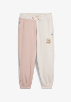 Bambino che indossa una tuta Puma a blocchi di colore beige e bianco con sneakers rosse, in piedi davanti a uno sfondo grigio uniforme.