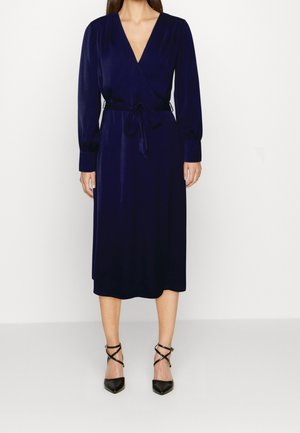 Robe portefeuille bleu marine avec des manches longues, une ceinture à la taille et un décolleté en V profond. Elle est fabriquée dans un tissu lisse et présente une coupe mi-longue. Portée avec des talons hauts noirs.