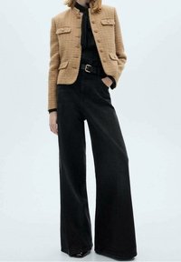 Personne portant une veste boutonnée texturée camel sur un chemisier noir avec nœud, accompagnée d'un pantalon noir taille haute à jambes larges et d'une ceinture noire.