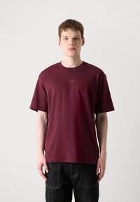Bordeauxfarbenes Baumwoll-T-Shirt mit Rundhalsausschnitt und kurzen Ärmeln. Dezentes, geprägtes "BOSS"-Logo auf der Vorderseite, kombiniert mit dunklen Jeanshosen.