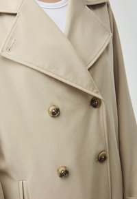 Beige dubbelbreasted jas met een brede revers, voorzien van grote bruine knopen en een gladde textuur. Zijzakdetail zichtbaar.