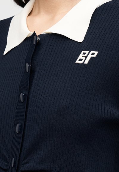 Ναυτικό ριγωτό ζακέτα με λευκό γιακά, με κουμπιά μπροστά και κεντημένο λογότυπο "BP" στο αριστερό μέρος.