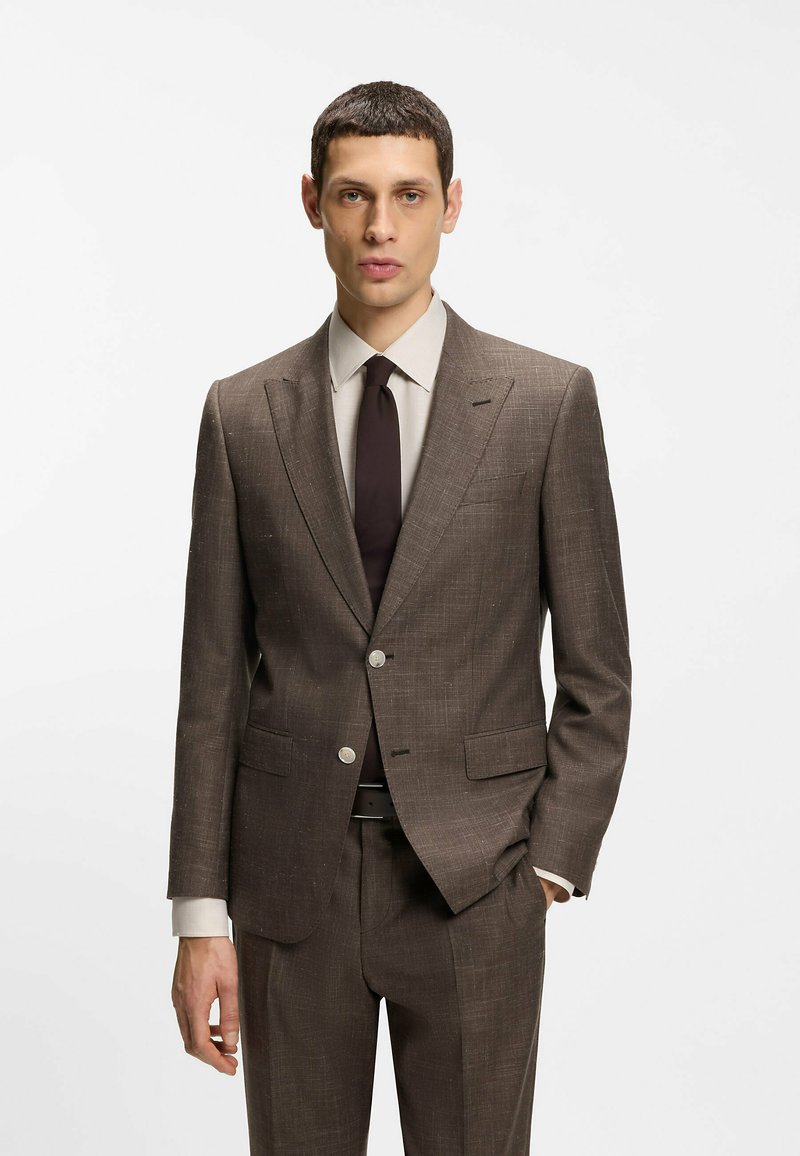 Homme portant un costume marron à carreaux, une chemise beige et une cravate marron foncé, debout avec une main dans la poche sur un fond blanc uni.