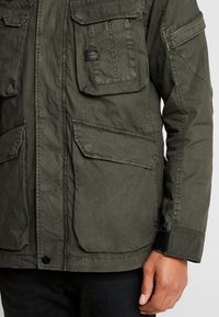 Olivgrön militärjacka med fyra framfickor, knapplåsning och justerbara ärmslut. Texturerat tyg med en avslappnad passform.