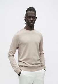 Bež dolg pulover z dolgimi rokavi iz mehke tkanine, z okroglim izrezom, rebrastimi manšetami in gladko teksturo. Oblečen s svetlimi hlačami.