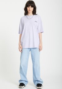 Licht lavendel oversized t-shirt met korte mouwen en een kleine grafische print op de borst, gecombineerd met lichtblauwe broek met brede pijpen en zwarte sneakers.
