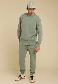 Ensemble sweat-shirt et pantalon vert clair en tissu doux avec des poignets côtelés. La casquette assortie arbore un logo. Les chaussures présentent des rayures noires et des accents beiges.