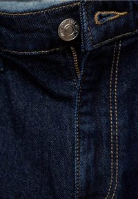 Jean en denim bleu foncé avec bouton en métal visible et fermeture éclair partiellement ouverte, présentant une couture contrastante jaune autour de la boutonnière.
