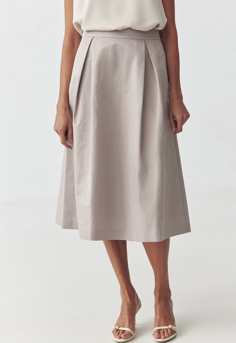 Jupe plissée grise pâle avec une texture lisse, de longueur genoux, dotée d'une taille et de poches latérales, associée à des sandales plates blanches.