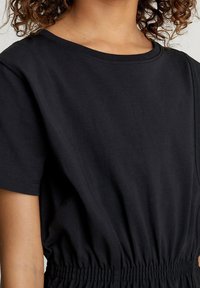 Musta puuvillainen paita, jossa on lyhyet hihat, pyöreä pääntie ja joustava vyötärönauha; sileä pinta ja hienovaraiset saumaustiedot.