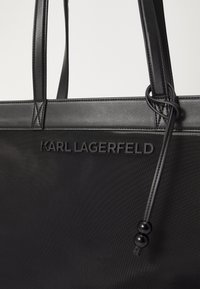 Black tote bag with textured nylon body, leather accents, and embossed "KARL LAGERFELD" logo. Features sturdy handles and a cinched closure.