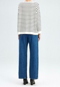 Person, der mit dem Rücken zur Kamera steht und ein gestreiftes Oberteil mit langen Ärmeln in weiß und schwarz, weit geschnittene blaue Jeans und schwarze Stiefel mit Absätzen trägt.