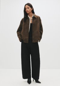 Rjava antilopska jakna s ovratnikom in sprednjimi gumbi, v kombinaciji z velikimi črnimi hlačami in črnimi koničastimi petkami na enobarvni beli podlagi.