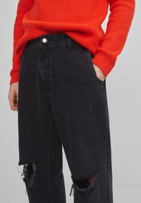 Persona vistiendo un suéter acanalado naranja brillante y jeans negros rasgados con agujeros en las rodillas, con una mano en el bolsillo frente a un fondo simple.