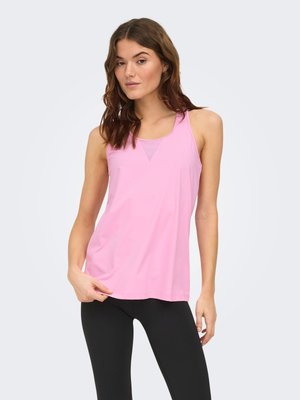 Femme aux cheveux bruns portant un haut de sport rose sans manches et un legging noir, debout devant un fond clair uni.