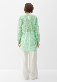 Donna con capelli corti castani, che indossa una blusa a maniche lunghe floreale verde menta e pantaloni bianchi a gamba larga, in piedi con le spalle rivolte verso di noi.