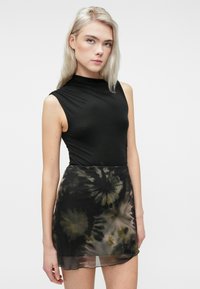 Mlada ženska z dolgimi blond lasmi, oblečena v brezrokavni črni top in mini krilo s črno tie-dye vzorcem, stoji ob enobarvni ozadju.