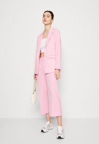 Blazer cintré rose clair et ensemble de pantalons raccourcis, associé à un haut blanc court et un sac à main blanc, rehaussé par des baskets grises et jaunes.