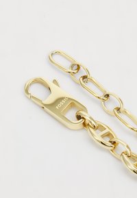 Cadena de metal dorado con eslabones ovalados y grandes eslabones circulares, con un cierre seguro marcado "FOSSIL". Superficie suave y brillante.