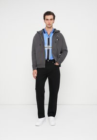 Szara bluza na zamek noszona na niebieskiej i białej koszulce z krótkim rękawem, w połączeniu z czarnymi spodniami i białymi trampkami. Prosty, casualowy wygląd.