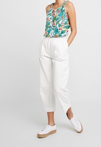 Nainen, jolla on yllään valkoiset korkeavyötäröiset housut, valkoiset korkeat tennarit ja olkaimeton trooppinen kukkakuosiset toppi, kädet taskuissa.