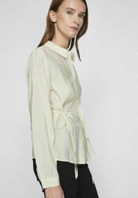 Blouse crème à manches longues avec un devant boutonné, détail de taille à nouer et tissu doux et texturé. Ajustée au niveau des poignets, coupe décontractée.