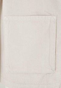 Gros plan d'une poche plaquée en tissu beige clair avec des coutures apparentes sur un vêtement.