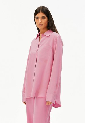 Frau mit langen, dunklen Haaren, die ein locker sitzendes rosa Hemd mit Knöpfen und passende rosa Hose trägt, vor weißem Hintergrund stehend.